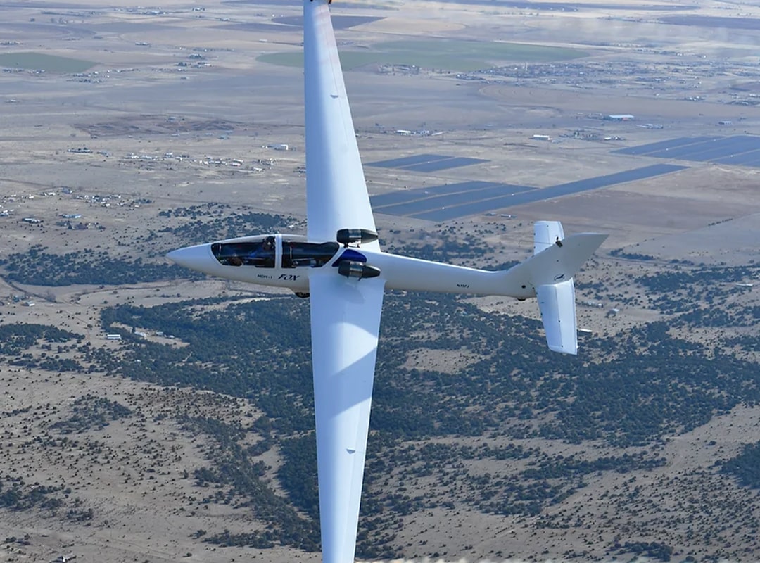 A sleek white Foxjet sailplane, piloted by Bob Carlton, is captured mid-flight performing an aerobatic maneuver against a backdrop of arid terrain and scattered vegetation. The aircraft is banked sharply, showcasing its long wingspan, twin jet engines, and aerodynamic design under clear skies.