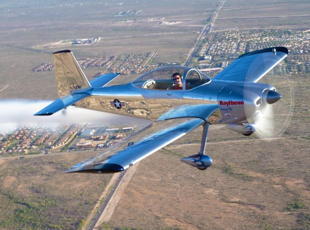 Polished silver RV-8 aircraft flown by Joe Shetterly in aerobatic flight over suburban landscape, featuring military insignia, Raytheon branding, and trailing smoke against clear skies.