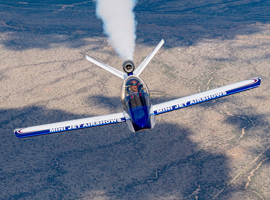 Tom Larkin flies his JSX003 SubSonex mini jet in an aerobatic airshow performance, trailing white smoke with "Mini Jet Airshows" displayed on the wings, against a rugged desert landscape.